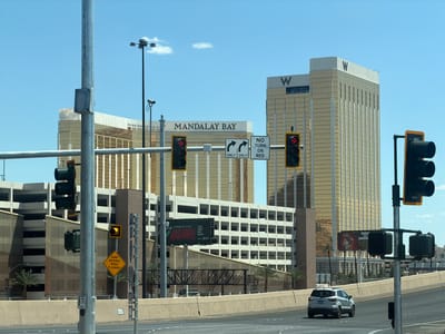 The image captures a cityscape in Paradise, Nevada, USA, featuring iconic landmarks such as the Mandalay Bay and the W Hotel. The scene is viewed from a road, with a silver SUV driving on it. Multiple traffic signals with red and yellow lights are visible, along with road signs including "RAMP METERED WHEN FLASHING", "ONLY", "NO TURN ON RED", and advertisements for "MAD APPLE CIRQUE DU SOLEIL" and "DINNER AND A SHOWDOWN TOURNAMENT OF KINGS". The weather appears to be sunny. The backdrop comprises high-rise buildings, a parking structure, and palm trees.