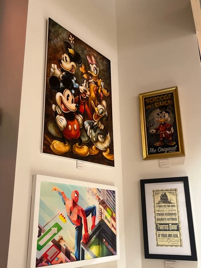 This image captures an interior wall display within a gallery or shop located in Chessy, France. The scene presents four framed artworks mounted on light-colored walls that form an inner corner.
On the upper left, a large vertical canvas displays a vibrant, dark-toned painting featuring classic Disney characters: Mickey Mouse, Minnie Mouse, Donald Duck, Daisy Duck, Goofy, and Pluto, rendered in a stylized, slightly aged aesthetic. Mickey Mouse is prominently positioned in the foreground, smiling. A small white informational label is affixed to the wall directly below this artwork.
To the right, on the adjacent wall, a smaller, vertically oriented artwork is housed in a golden frame. This piece is a poster-style print depicting Scrooge McDuck, with visible text including "SCROOGE MCDUCK," "Crucial Since 1947," and "The Original Tycoon." A small white informational label is present below its frame.
Below the large Disney character painting, on the lower left, a medium-sized horizontal graphic art print is framed in white. This artwork depicts Spider-Man in a geometric, polygonal style, perched on a building against a stylized urban backdrop of various colored buildings and architectural elements. A small white informational label is positioned beneath this artwork.
Directly below the framed Scrooge McDuck print, on the lower right, a medium-sized vertical print is enclosed in a black frame with a white mat. This print resembles a vintage poster titled "PHANTOM MANOR," detailing "STRANGE OCCURRENCES AND ENIGMATIC HISTORIES" and advising that visits are "AT THEIR OWN RISK." A small white informational label is affixed beneath this framed piece.
The walls are uniformly light-colored, and the lighting is consistent throughout the displayed area, indicating an intentional exhibition setup.