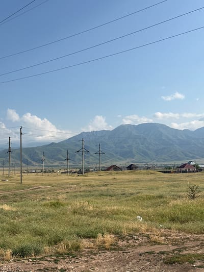 The image depicts a rural landscape in the vicinity of Sary-Moldoyev Rural District, Kazakhstan, under a clear blue sky with scattered white clouds. The foreground is a dirt path transitioning into a field of dry grass with patches of green vegetation. Several tall, wooden utility poles with power lines stretch across the middle ground, interspersed with some low-lying structures and houses with red and brown roofs. In the background, rolling, green hills form a mountain range, with some darker shadows suggesting valleys and contours. The overall impression is of an undeveloped, agricultural or semi-rural area.