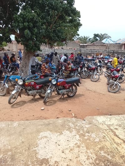 The image shows a group of people gathered around motorcycles in an outdoor, open-air area. There are approximately 20 motorcycles, mostly red and blue in color, parked in a row. Several people are sitting on the motorcycles while others stand around. The scene is likely in front of a building, as a brick wall is visible in the background. The ground is dusty and uneven, with a concrete area in the foreground. The setting is a residential area in Mukaiya, Nigeria. The large tree in the center of the image casts a shadow over the motorcycles and people. The overall scene is one of casual gathering and socializing.