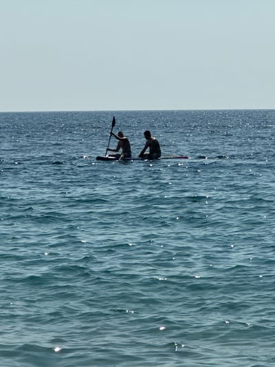 The image depicts two individuals on paddleboards on a body of water. The water is a deep blue with choppy waves, suggesting a slightly breezy day. Sunlight glints off the water's surface, indicating it is likely daytime, perhaps late morning or afternoon. The sky is a pale, hazy blue, suggesting a clear but possibly overcast day.
The first person, positioned slightly higher in the frame and further back, is a person of darker complexion, crouched low on their paddleboard. They appear to be in motion. The second person, closer and lower in the frame, is also on a paddleboard and is leaning forward, seemingly in the process of paddling or maintaining balance. Both individuals are dressed in dark clothing.
The scene is set on a vast expanse of water, with no land or other distinguishing features visible beyond the two paddleboarders. The focus is on the dynamic interaction between the individuals and the water. The overall impression is one of active recreation and skill on the water. The location context of Mirište, Montenegro, implies a coastal or lake setting within that region. There is no visible text in the image.
