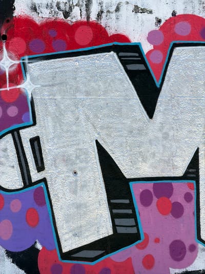 This media file is a close-up, detailed photograph of graffiti art on a textured concrete surface, likely an exterior wall. The graffiti features a large, stylized silver letter or symbol with a bold black outline accented by a thin blue line. The artwork is embellished with a pattern of red and purple circular shapes, resembling polka dots, in various sizes, against a lavender background. The underlying concrete wall shows signs of wear and tear, with patches of peeling paint and a rough, uneven texture.

The lighting suggests it is daytime, with clear illumination that highlights the texture of the wall and the vibrancy of the paint. There are no people or other objects visible in the frame, focusing solely on the abstract graffiti art. The style of the graffiti, with its sharp lines and vibrant colors, is characteristic of urban street art. The location context provided, l'Hospitalet de Llobregat, Spain, suggests this artwork is part of the city's urban landscape. There is no visible text within the graffiti or on the wall itself. The overall impression is a dynamic and colorful piece of street art contrasted with the raw, weathered texture of the wall.