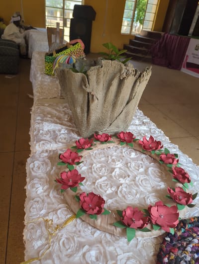 The image displays a table covered with a textured, white cloth adorned with embossed rosettes. On the table, there is a decorative circular wreath made of fabric flowers in shades of red and green. The wreath is adorned with small, sparkly embellishments at the center of each flower. A gold ribbon is attached to the wreath, trailing down the side of the table. To the left of the wreath, a rough-textured, beige object that resembles a piece of driftwood or sculpted concrete stands upright, with a small green plant growing from its opening. Further to the left, a black speaker is visible. The background shows a window with sunlight streaming through, suggesting daytime. In the distance, a banner with text is partially visible. The overall setting appears to be indoors, possibly a community space or exhibition. The focus of the image is on the decorative elements displayed on the table.