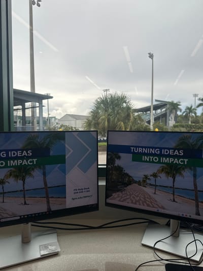 The image presents an interior view looking out through a large window, with two computer monitors positioned on a counter in the foreground. The scene is located at Florida Gulf Coast University (FGCU), near Miromar Lakes, United States, as indicated by "fgcu.edu/IMPACT" displayed on the monitors.

In the immediate foreground, two identical flat-panel Dell monitors with white stands are situated on a light-colored, speckled countertop. Black cables are visible behind and around the monitor bases. A small white card or paper rests on the base of the left monitor. Both monitors display a graphic featuring palm trees lining a waterfront with a paved walkway, beneath a blue and green banner. Text on the monitors includes "TURNING IDEAS INTO IMPACT," "ITS Help Desk," and the phone number "239-590-1188," along with "fgcu.edu/IMPACT."

Beyond the window, an overcast, light gray sky dominates the upper portion of the frame. The outdoor environment features several palm trees. To the far left, a portion of a covered structure or pavilion with support columns is visible. In the midground and background, multiple tall poles equipped with stadium lighting fixtures are prominent. A large stadium or bleacher structure with visible dark seating and a metal framework is present on the right side of the view. A white, rectangular building and other less defined structures are discernible in the distance. No individuals are visible within the frame.