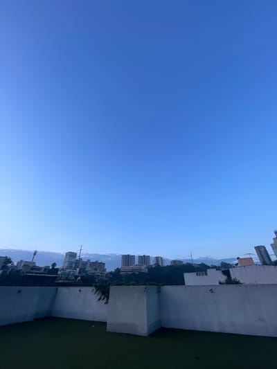 This is a full-frame, eye-level, vertical shot taken from a rooftop in Huixquilucan de Degollado, Mexico, looking out over a cityscape. The foreground features a white, L-shaped wall enclosing a patch of dark green artificial turf. In the background, a dense collection of buildings rises from a tree-covered hillside. Among these structures are several high-rise apartment buildings and smaller residential buildings, with some appearing to be under construction. The sky above is a clear, gradient blue, transitioning from a lighter shade near the horizon to a deeper hue at the top of the frame. The lighting suggests it is either early morning or late evening. There are no people visible in the scene.