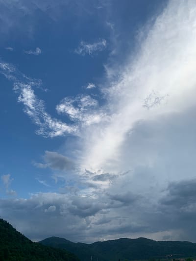 This is a vertically oriented, slightly tilted, wide-angle shot of a dramatic sky over a mountainous landscape. The sky is filled with a dynamic mix of clouds. On the left, there is a clear, bright blue sky with wispy white cirrus clouds scattered across it. As the eye moves to the right, the clouds become denser and darker, transitioning into heavy cumulonimbus formations that appear to be either building or dissipating. The lighting suggests it is daytime, likely late afternoon, as there is a mix of bright sunlight filtering through some clouds and dark shadows cast by others.
The right side of the image features a dark, densely forested mountainside. The slope of the mountain is steep, and the trees are a deep green. Along the very edge of the mountain, faint signs of human habitation are visible, appearing as scattered lights or small structures, suggesting a village or town nestled on the mountainside. The overall mood of the image is awe-inspiring and perhaps a little foreboding, given the dramatic cloud formations and the imposing presence of the mountain. The location context provided is Khust, Ukraine, which aligns with the mountainous terrain. There is no discernible text in the image.