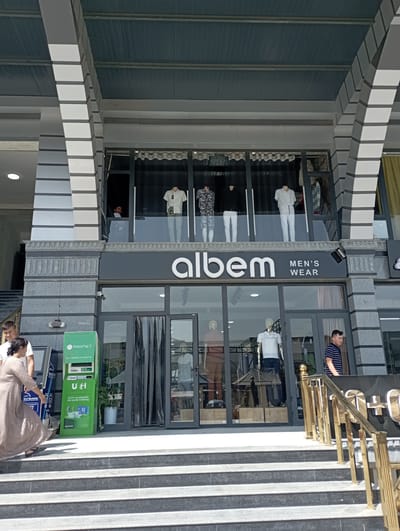 This image depicts the exterior of "albem MEN'S WEAR" in Termez, Uzbekistan. The storefront features a large glass facade on the ground floor, with mannequins displaying men's clothing visible inside. Above the ground floor, there is another level with mannequins in the windows. A green "PAYNET" payment terminal is situated to the left of the entrance, with a woman in a light-colored dress and a man in a white t-shirt standing near it. Steps lead up to the entrance of the store. The building has a modern architectural style with prominent gray and white columns. The overall scene appears to be a sunny day.