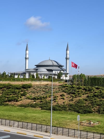 The image shows a mosque with two minarets and a large dome, situated in a landscaped area. A Turkish flag is visible flying on a pole to the right of the mosque. In the foreground, there is a grassy slope with dense green bushes and a paved walkway with a black metal fence. The sky is clear blue with a few wispy clouds. The overall scene suggests a sunny day in a modern urban or suburban setting. The architectural style of the mosque indicates a contemporary design. The location is identified as Arnavutköy, Turkey.
