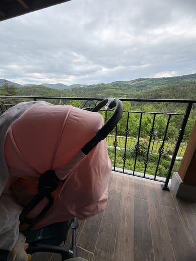 A baby stroller with a pink canopy sits on a tiled balcony. The balcony has a black metal railing. Beyond the railing, a landscape of green trees and distant mountains is visible under a cloudy sky. The location is Кирково, Bulgaria.
