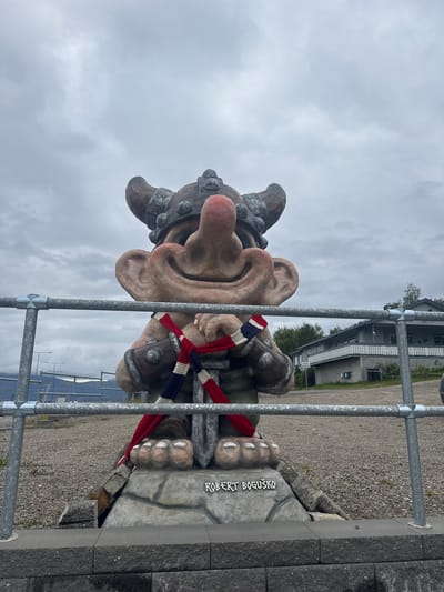 A large, grey-skinned troll statue stands outdoors. The troll has a large nose, prominent ears, and is wearing a metal helmet with horns. It has a red, white, and blue striped scarf around its neck. The statue is situated on a stone base. In front of the statue is a metal railing. In the background, there is a building with a light-colored facade, trees, and a cloudy sky. The ground appears to be covered in gravel. A name, "ROBERT BOGUŠKO", is inscribed on the base of the statue. The location is identified as Storjord i Tysfjord, Norway.