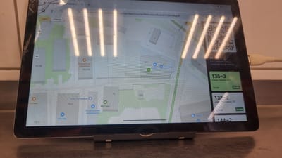 The image shows a tablet screen displaying a map interface with superimposed order information. The map indicates a location in Megion, Russia. On the screen, a QR code is visible, with text instructing users to scan it to join the queue. Below the QR code, there are order details, including order numbers, street names, and estimated waiting times. For example, order "135-3" is located at "улица Ленина, 4/1" and is marked as "Готов" (Ready) with a 22-minute waiting time. Another order, "138-2", is at "улица Кузьмина, 10" with a 24-minute waiting time. The tablet is connected to a power source via a USB cable. The environment appears to be a countertop or table. The screen has some smudges and glare from lighting.