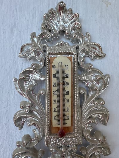 A close-up, high-angle, eye-level shot captures an ornate, silver-colored thermometer mounted on a textured, white wall. The thermometer, encased in a decorative frame with floral and scrolling motifs, displays a temperature reading of approximately 25 degrees Celsius. The thermometer's glass tube contains a red liquid that has risen to just below the 30-degree mark, with clear markings for every 10 degrees and finer divisions in between. The wall's surface exhibits a subtle, uneven texture, suggesting it might be painted wood or plaster. The overall scene is static and devoid of any human presence or activity, presenting a solitary artifact against a neutral backdrop. The location is identified as Peremyshliany, Ukraine.