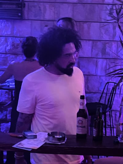 The image depicts a dimly lit indoor scene, possibly a bar or restaurant, bathed in purple ambient light. In the foreground, a man with curly dark hair and a beard, wearing a light-colored t-shirt, stands at a dark wooden counter. He is looking to his left. On the counter, there are two beer bottles, one with a white label and the other dark green, an ashtray with a cigarette butt, and what appears to be a pack of cigarettes and some papers. In the background, a woman with dark hair is visible from the back, wearing a black dress. Another person's head is partially visible behind the man in the foreground. The wall behind them is made of textured stone blocks. The scene suggests a relaxed, social atmosphere.