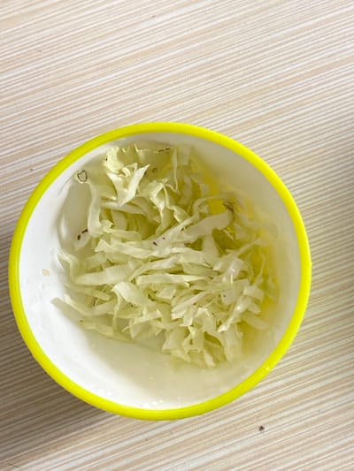 An overhead shot captures a white bowl with a bright yellow rim, filled with finely shredded light green cabbage. The bowl is positioned towards the right side of the frame, resting on a light beige or tan surface that features a subtle, repetitive diagonal striped pattern. The cabbage is uniformly cut, appearing fresh and slightly moist, with a few small, dark specks interspersed among the shreds. No individuals are present in the image, and no specific activity is taking place; the scene depicts a static arrangement of food. The setting suggests an indoor environment, possibly a kitchen or dining table, located in Bekasi Regency, Indonesia. The lighting is bright and even, illuminating the contents of the bowl. There is no visible text in the image.