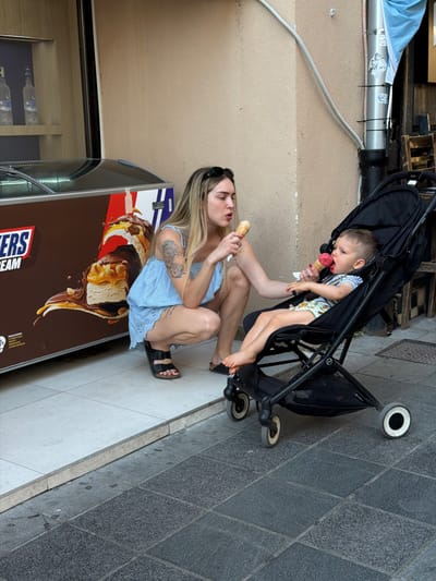 In Sarajevo, Bosnia and Herzegovina, a woman is crouching on the sidewalk in front of an ice cream freezer displaying an advertisement for Snickers ice cream. She is wearing a light blue dress and black sandals, with sunglasses pushed up on her head. The woman is holding an ice cream cone and offering it to a baby sitting in a black stroller. The baby, wearing a striped shirt and patterned shorts, is also holding an ice cream cone. The scene takes place on a street with tiled pavement and a concrete wall in the background.