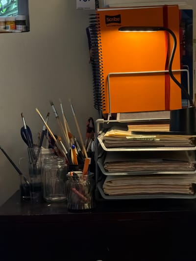 An orange notebook with a black spiral binding sits under a gooseneck lamp. Adjacent, a file organizer holds three filled folders. Below, clear jars and a black mesh container hold a collection of paint brushes. The scene is dimly lit.