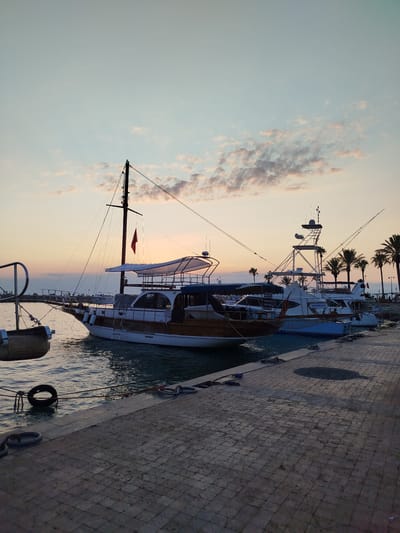 The image depicts a tranquil harbor scene at dusk in Manavgat, Turkey. Several boats are docked along a paved walkway. In the foreground, the textured paving stones lead to the water's edge, where various ropes and a tire fender are visible. The water is calm, reflecting the soft light of the setting sun.
The most prominent boat in the midground is a wooden-hulled vessel with a white canopy and a single mast flying a red flag. To its right, a more modern, white yacht with multiple decks and antennae is docked. Further down the pier, more boats and palm trees are silhouetted against the fading light. The sky is a gradient of pale blue and soft orange, with scattered clouds tinged with pink and purple. A few distant figures can be seen strolling along the waterfront in the background, suggesting a public space. The overall atmosphere is peaceful and serene, characteristic of a harbor at the end of the day.