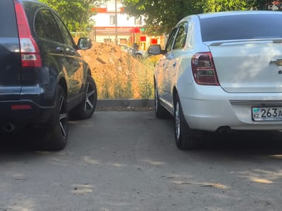 This image captures two cars parked side-by-side on a paved surface, likely a parking lot or a street. The car on the left is a dark-colored SUV, seen from the rear passenger side. Its taillight and large rear wheel with a distinctive rim are visible. The car on the right is a white sedan, also viewed from the rear passenger side. It has a spoiler on the trunk and a license plate that reads "KZ 263A" with additional text below.
In the background, there's a mound of earth, suggesting a construction or landscaping site. Beyond that, a building with white and red sections is visible, along with trees that provide shade, indicating an outdoor setting. The lighting suggests it's daytime, possibly late afternoon given the shadows cast on the ground. The weather appears clear. The presence of the license plate with "KZ" clearly indicates a location in Kazakhstan, aligning with the provided location context of Abay, Kazakhstan. No people are visible in the image, and the cars appear to be stationary. The scene conveys a sense of everyday life, with vehicles parked in a common area.