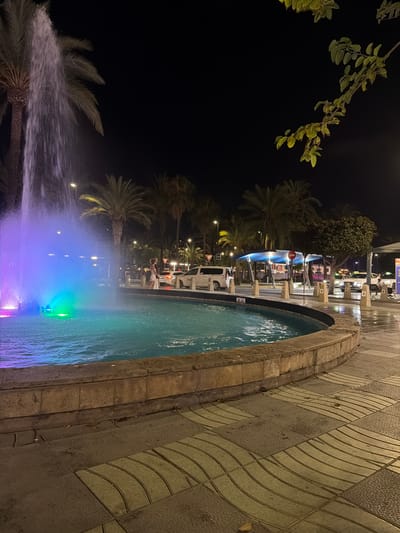 The image captures a nighttime scene at a fountain in Sant Antoni de Portmany, Spain. The fountain is illuminated with vibrant blue and purple lights, casting a colorful glow on the water and the surrounding area. Palm trees, silhouetted against the dark sky, are visible in the background, adding to the tropical ambiance. The ground is paved with textured tiles, and a stone border surrounds the fountain. A few people are present in the scene, some standing near the fountain and others in the background, possibly dining or socializing at outdoor seating areas. The time of day is clearly night, suggested by the dark sky and artificial lighting. The overall mood appears to be relaxed and festive, characteristic of a pleasant evening in a tourist destination.