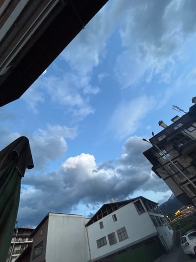 The image captures a low-angle view looking upwards towards a mixed sky, framed by architectural elements and an umbrella. The photograph is rotated, giving the impression that the surrounding structures are leaning.
The sky features patches of clear, light blue interspersed with a variety of clouds. To the left and center, lighter, wispy clouds are visible, while towards the right and upper areas, the clouds are denser, darker, and more grey, suggesting variable weather conditions.
Several buildings of different styles form the foreground and midground. In the upper section, an older, multi-story building with multiple windows and balconies is prominent, extending into the frame with what appears to be a streetlight. On the right side, a more modern, predominantly white building with several windows is visible. In the background, between the buildings, a dark mountain range can be faintly discerned. A portion of a white car is visible in the upper right corner, near a building.
In the lower left of the frame, a dark roof edge or awning is present. In the lower right, a large, dark green or black patio umbrella, in a folded state, partially enters the view.
There is no specific human activity evident in the static scene. The time of day appears to be daylight, likely during the mid-afternoon, given the sky's illumination and varying cloud cover. A distinctive detail is the word "CASINO" visible on a sign attached to one of the buildings, located below the modern white building. The combination of buildings, mountains, and the casino sign suggests an urban area within a mountainous resort environment, consistent with Bansko, Bulgaria.