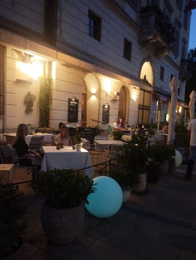 The image depicts an outdoor dining area of a restaurant in Lugano, Switzerland, during the evening. The facade of a building is visible, featuring a sign with the word "ristorante" and windows illuminated from within. Several tables are set up with white tablecloths, some occupied by patrons. Two women are seated at a table in the foreground, engaged in conversation, with drinks and place settings on the table. Another group of people are seated at tables further back, appearing to dine or socialize. Large potted plants and decorative elements are placed around the seating area. A large, illuminated blue sphere is visible on the right side of the image. The ground is paved with dark tiles. The overall ambiance suggests a relaxed evening at a European restaurant.