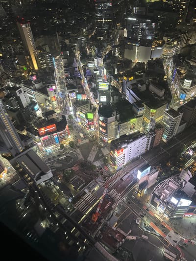 The image captures an aerial nighttime view of Shibuya, Japan, characterized by densely packed buildings illuminated with an array of lights. A significant pedestrian scramble crossing, crowded with people, is prominently featured. Major buildings and structures include train tracks and stations, contributing to the area's transportation infrastructure. The general environment suggests a bustling urban atmosphere at night.