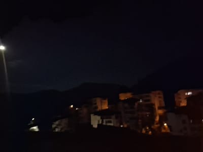 This is a nighttime, slightly blurred, horizontal medium shot of a town or city nestled at the base of mountains. A single streetlamp is visible on the far left, its light illuminating the immediate foreground. The background features dark, silhouetted mountains against a deep blue-black sky. In the middle ground, several multi-story buildings are visible, with some windows illuminated, suggesting an urban or semi-urban setting. The lights from the buildings are scattered and appear as small yellow or white dots. The overall impression is one of a quiet, dimly lit night scene. The image is likely taken in Smolyan, Bulgaria, given its mountainous terrain and common architectural style of buildings seen in the region.