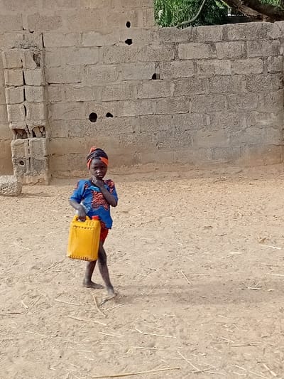 The image displays a young child standing outdoors, holding a yellow jerrycan. The child is positioned slightly to the right of the center of the frame and is looking directly at the camera. They appear to be a toddler or a very young child, dressed in a long-sleeved shirt or dress with a blue and red patterned fabric, and a dark head covering. Their feet are bare.
The child is holding a bright yellow plastic jerrycan or container with both hands, positioned in front of their body. The jerrycan has a ribbed texture on its sides.
The setting appears to be a dry, open area. To the left of the child, there is a wall constructed from light-colored, possibly concrete or mud, blocks. The wall shows signs of wear, with some cracks, uneven surfaces, and several circular holes or openings at various heights. Towards the bottom left of the wall, some blocks appear damaged or missing. Behind the upper left section of the wall, some green foliage or leaves are visible, suggesting the presence of a tree or plant.
The ground beneath the child and extending into the foreground and background is a light brown or sandy color, appearing dry and dusty. Scattered throughout the ground are light-colored pieces of dry grass or straw.
The lighting suggests it is daytime, with bright, even illumination. There is no visible text in the image.