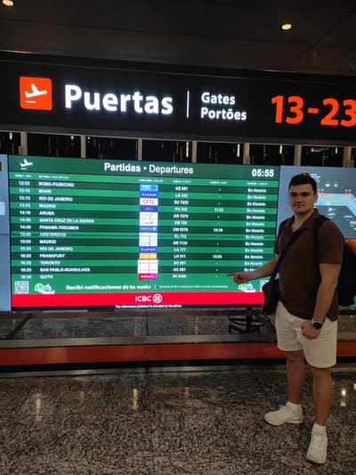 In this medium shot, a young man stands in front of a large digital departure board at Aeropuerto Internacional Ezeiza, Argentina. The board displays flight information with columns for time, destination, airline, flight number, gate, and status. The man, wearing a brown polo shirt, white shorts, and white sneakers, is pointing towards the board with his left hand, which has a watch on his wrist. He carries a black shoulder bag. The departure board is illuminated with a bright green and red section. The airport environment is visible with a granite-patterned floor and a reddish-orange horizontal accent above the floor. The man is positioned to the right of the departure board, and the overall lighting suggests an indoor airport setting.