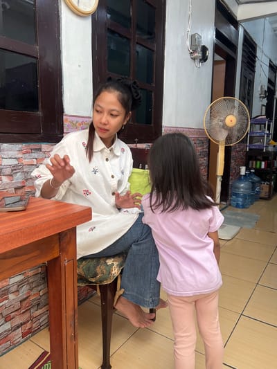 In the city of Yogyakarta, Indonesia, a young woman and a young girl are engaged in conversation in what appears to be a home. The woman, with her hair styled in a large bun, is seated on a chair and wearing a white shirt adorned with floral embroidery and blue jeans. She gestures with her hands as she speaks to the girl, who stands facing away from the camera. The girl is dressed in a light pink t-shirt and pink leggings.
The environment suggests a living space with a wooden table in the foreground. Behind the woman, a patterned wall can be seen, and further back, a yellow oscillating fan stands near large water dispenser bottles. The room has tiled flooring and wooden window frames. A clock is visible on the wall above the woman. The overall scene is casual, implying a domestic setting within Yogyakarta.
