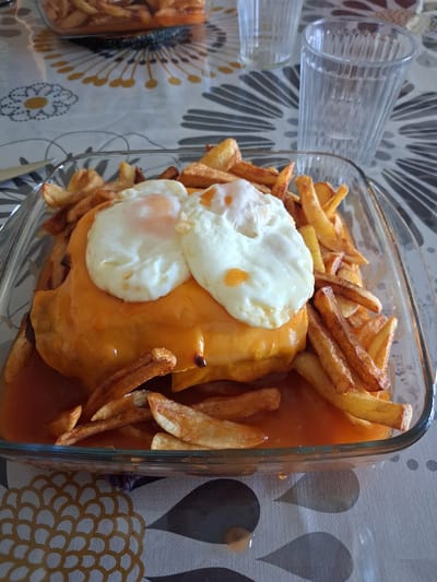 A Francesinha, a Portuguese sandwich, sits in a clear glass baking dish. Two fried eggs top the sandwich, which is covered in melted cheese. The sandwich is surrounded by french fries and a reddish-brown sauce. To the left of the dish are two clear drinking glasses, and to the bottom left is another container with french fries. The dish is placed on a tablecloth with a geometric pattern of gray, white, and yellow. The location is Figueira da Foz, Portugal.