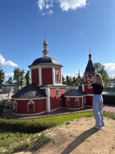 A young woman with blonde hair, wearing a black t-shirt and light blue jeans, stands on a sandy slope, taking a photograph of a vibrant red church with a dark roof and gold-tipped domes. The church, which appears to be part of the Suzdal Kremlin in Suzdal, Russia, features white trim and architectural details, including arched windows. Behind the main church, a bell tower with a similar color scheme and a cross on top is visible, along with other buildings and trees in the background. The sky is a bright blue with scattered white clouds, and the sun casts a long shadow of the woman on the ground. The foreground shows green grass and a hedge.