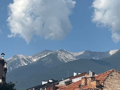 The image captures a serene mountain landscape above the town of Bansko, Bulgaria. In the foreground, the terracotta-tiled rooftops of several buildings are visible, some adorned with white chimneys. A portion of a building with a white facade and a brown tiled roof is partially seen on the far left. Beyond the rooftops, a dense, dark green forest blankets the lower slopes of the majestic mountains. The upper reaches of these mountains are characterized by rugged, rocky terrain with patches of snow, suggesting a high altitude. The sky above is a vibrant, clear blue, punctuated by fluffy white clouds. The overall impression is one of a picturesque vista combining urban elements with the grandeur of nature.