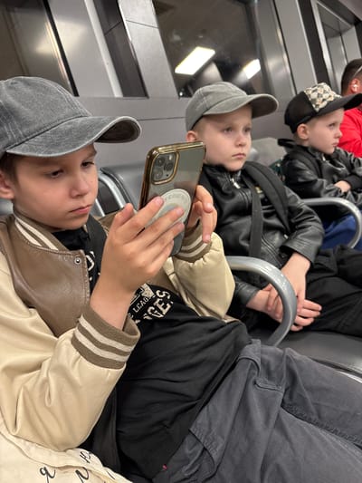 In Bol'shie Kabany, Russia, a group of three young boys and a partial adult are seated in an indoor public waiting area. The foreground features a pre-teen boy, wearing a grey baseball cap, a brown and off-white varsity-style jacket with striped cuffs, a black t-shirt with partially visible white text, and dark grey pants. He is actively engaged with a gold-colored smartphone, holding it with both hands and looking down at its screen. A circular sticker displaying an emblem and Cyrillic text is affixed to the back of the phone's case.
To his right, a younger boy, approximately 5-7 years old, also wears a grey baseball cap and a black faux leather jacket with visible backpack straps, paired with dark pants. He sits upright, observing something outside the frame to his right, with a neutral expression. Adjacent to him, a third boy of similar age wears a black baseball cap with a checkered pattern on the front and a black faux leather jacket over blue pants, with a backpack strap also visible. He mirrors the gaze of the second boy, looking towards the right.
The far right of the frame shows the upper portion of an adult male, clad in a red garment, with a white earbud visible in his right ear. All individuals are seated on grey, metallic-framed public seating. The environment consists of modern, light-colored and grey architectural panels on the walls, some exhibiting reflections, and overhead rectangular light fixtures contributing to a bright, artificial illumination.