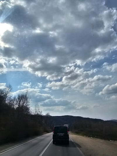 The image depicts a daytime, forward-looking view from a moving vehicle on a two-lane asphalt road in Bălteni, Romania. A dark-colored minivan, possibly black or dark blue, travels directly ahead in the same lane. The minivan's rear license plate is partially visible, displaying "RO", indicating Romanian registration. Its taillights are unilluminated. Further in the distance, a portion of a larger, lighter-colored vehicle is discernible on the right side of the road. No individuals are visible.
To the left of the road, a steep embankment or hillside rises, densely covered with bare deciduous trees and dark undergrowth. The right side of the road features a dirt shoulder. The background is formed by a range of hills or low mountains heavily covered in dark vegetation. The sky occupies the upper portion of the frame, characterized by prominent grey clouds interspersed with patches of blue sky. Direct sunlight, with visible rays, emanates from the upper-left quadrant.