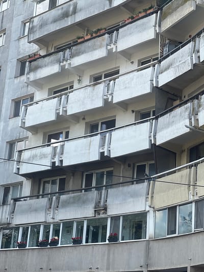 This is a low-angle, full shot of a Brutalist-style apartment building in Iași, Romania. The building features multiple levels of protruding concrete balconies with white awnings. These balconies are arranged in a staggered pattern, creating a visually dynamic facade. Many of the balconies have small planters with red flowers, adding touches of color to the otherwise grey concrete structure. The windows are a mix of dark-paned and reflective glass, with some showing glimpses of the sky and trees reflecting on their surfaces. The overall impression is one of architectural complexity and a somewhat stark urban environment. The time of day appears to be overcast, as there are no strong shadows. No people are visible in the image, and the scene suggests a quiet, residential setting. There is no visible text.