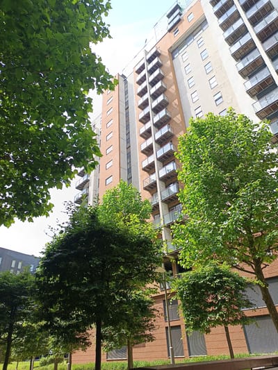 This is a low-angle, full-shot, outdoor photograph taken in Manchester, United Kingdom. The primary subject is a tall, modern apartment building with multiple stories, balconies with glass railings, and a mix of brick and concrete facades. Green trees, with lush foliage, are prominent in the foreground and mid-ground, partially obscuring the building and creating a contrast between the urban architecture and the natural elements. The sky above is mostly clear with some diffused sunlight. The image appears to be taken during daylight hours on a clear day.