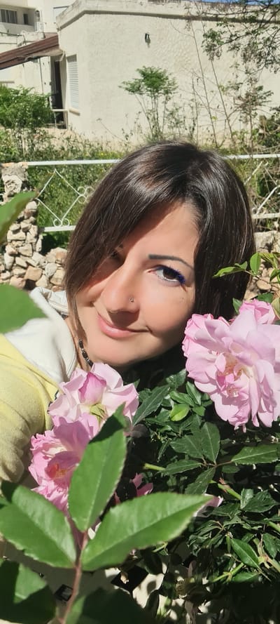 A medium shot depicts a smiling woman with dark brown hair. She is wearing blue eyeliner and has a nose piercing. Her clothing consists of a yellow top with a white collar. She is partially surrounded by pink roses and green foliage. A stone wall and white iron fence are visible in the background, along with a white building and some green trees. The location is likely Nahariyya, Israel.