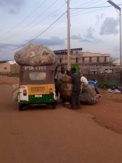 In Katon Rikkos, Nigeria, two men are loading large bags of what appears to be waste or recyclable materials onto a three-wheeled vehicle, commonly known as an auto-rickshaw or "Keke Napep." The auto-rickshaw is overloaded with a massive mound of bagged materials strapped to its roof. The vehicle itself is painted white, yellow, and green, with the license plate "PXH 544 VZ" visible. The number "PTA-8786" is also displayed on the back.
One man, wearing a dark beanie and a dark jacket, is actively engaged in lifting and positioning a large bag. The other man, dressed in a light-colored shirt, stands behind him, also involved in the loading process. In the background, a building under construction is visible, with scaffolding and exposed structural elements. The sky is overcast with dramatic clouds, suggesting either early morning or late afternoon. The ground is a reddish-brown dirt road. The scene conveys a sense of hard work and manual labor, likely related to waste management or recycling in the area.
