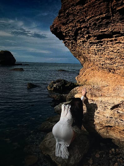 A person wearing a white dress is kneeling on rocks by the sea, their hands pressed against a large, textured rock formation. The person's back is to the camera, with long dark hair flowing down. The sea is dark blue and appears choppy, with several rocks visible in the water. The sky is partly cloudy with a bright blue hue. The location is identified as Chotěšov, Czechia.