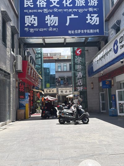 This daytime photograph captures a commercial street scene in Lhasa, China, specifically at No. 39 Yutuo Road, as indicated by visible signage. The perspective looks down a paved street lined with multi-story buildings on both sides. The bright sunlight casts sharp shadows, suggesting clear weather.
On the left, a large blue banner, featuring both Chinese and Tibetan script, advertises a "Folk Culture Tourism Shopping Plaza." Below it, an air conditioning unit is mounted on the building. The right side of the street features several parked motorbikes or scooters, some with white helmets. A few people are visible near these vehicles, with another person under an umbrella further down the street.
The buildings display various signs, including an overhead banner partially showing "Chin" (likely part of "China"). A prominent red LED sign above a banking facility indicates "24 Hours Self-Service Banking" in both English and Chinese. Another sign identifies "Yucan Hotel 3F." Other businesses advertised include "Demulin Sand Local Specialty Store," with names and phone numbers in both Chinese and Tibetan scripts. A red LED sign on the ground reads "源" (Source/Origin). The pervasive use of both Chinese and Tibetan languages serves as a strong location cue.
