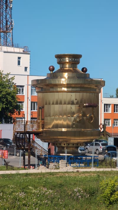 This image captures a large, ornate, brass-colored object resembling a samovar, placed outdoors in an urban or semi-urban setting. The object is positioned vertically, with its base towards the bottom of the frame and its top extending upwards. It appears to be a decorative installation or sculpture.
Surrounding the samovar, there are several buildings, including a white and orange multi-story building, and a building with white walls and orange accents. In the background, there are parked cars, including a silver SUV and a dark-colored van or SUV. A blue metal structure, possibly shelving or a temporary construction element, is also visible.
To the right of the samovar, there is a grassy area with some small pink wildflowers and green foliage. A pathway or sidewalk runs along the edge of the grass. In the foreground, there's a glimpse of what might be a construction site with metal scaffolding or framework.
The scene appears to be set during the daytime, under a clear blue sky. The lighting suggests it might be late afternoon or early evening, given the warm reflections on the brass. There are a few people visible in the lower part of the image, near the decorative metalwork in front of the samovar, one appearing to be wearing a teal shirt. One person seems to be working near the metal structure.
A few pieces of text are faintly visible on a red and yellow sign in the lower left, but the text is too blurry to be read. The overall impression is of a public space with a unique, oversized art piece. The location context points to Bor, Russia.
