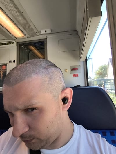 The image shows a close-up of a person from the shoulders up, inside what appears to be a train carriage. The person has a shaved head and is wearing a white t-shirt and black wireless earbuds. They are looking down and to the right, with their face partially obscured.

The interior of the train is visible, with a fluorescent light fixture on the ceiling and a window showing a blurred outdoor scene with trees and buildings. A digital display on the train wall shows "126 16:35". There is a red emergency pull handle on the wall. The seat upholstery is blue and white checkered.

The person's head is positioned in the foreground, dominating the frame. The focus is sharp on the earbud and the side of the face. The overall lighting suggests it is daytime.

While the image is taken inside a train, there is no specific visual information within the image itself to confirm the location as Zempin, Germany. The digital display showing "126 16:35" does not provide geographical context.