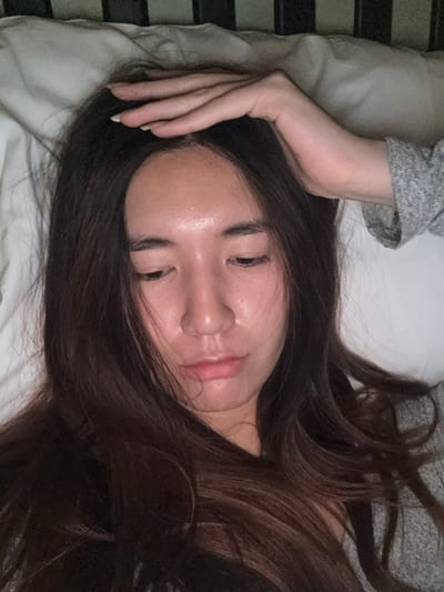 A young adult female is depicted in a close-up, overhead shot, lying on a bed indoors. She possesses long, dark brown hair, which frames her face and spreads across the white pillow. Her face is angled slightly downward and to the viewer's left, with eyes cast low or partially closed, and a neutral, slightly pensive expression. Her skin appears luminous. Her right arm is raised, with her hand resting on the top of her head and forehead, fingers subtly splayed. She wears a light gray, textured long-sleeved garment on her visible arm. The bedding consists of a white pillow and sheets. A dark, indistinct horizontal structure, possibly a bed frame or headboard, is visible above her head against the light background. The lighting is dim, suggesting an indoor setting. The image does not contain any visual information to determine a specific location within the city of Le Havre, France, or any other city.