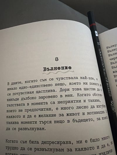 The image shows an open book with text in Bulgarian and a black pencil with "SUPERIOR QUALITY BLACK LEAD PENCIL" printed on it. The book is open to page 8, which has the title "Вълнение" (Excitement). The text on the page discusses feelings of happiness and the desire to live and be motivated, especially during difficult times. There are no people or animals visible in the image. The environment appears to be indoors, with a soft gray surface behind the book, suggesting it might be on a couch or bed. The lighting is dim, casting shadows on the pages. The image does not provide any information about the location in the city of Svilengrad, Bulgaria.