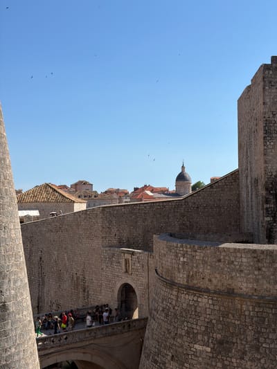 The image shows a view of the historic city walls of Dubrovnik, Croatia, on a clear, sunny day. In the foreground, a large, weathered stone wall and a cylindrical tower on the left dominate the scene, indicating the fortifications. To the right, a curved section of the wall extends into the frame. A stone bridge with decorative railings spans an archway, and a group of tourists are walking across it. The bridge leads to an arched entrance in the wall. In the middle ground, the red-tiled roofs of buildings in the old town are visible, and a distinctive domed building, likely a church or cathedral, rises above them. The sky is a bright, clear blue, with several small birds flying in the distance. The overall impression is one of historical significance and a popular tourist destination.