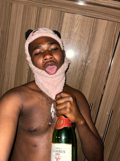 A shirtless male individual with dark skin is depicted, wearing a pink, fuzzy head covering resembling a panda balaclava, which extends over his head, ears, chin, and neck. He holds a green glass bottle labeled "ST. LAUREN RED" with a red cap in his left hand. His tongue is extended from his mouth. A white, viscous substance is visible on his chest, dripping from below the head covering. The background consists of vertical light brown wood-grained cabinetry or paneling, with a reflective horizontal strip visible near the top. The specific location within Muchiya, Nigeria, cannot be determined from the visual evidence provided in the image.