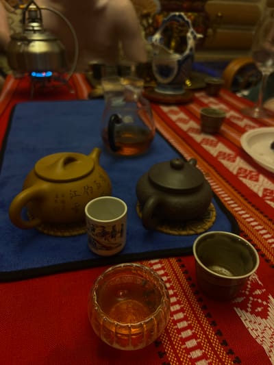 An indoor scene showcases a tea preparation and serving setup on a table. The table is covered by a vibrant red and white patterned tablecloth, featuring geometric and possibly floral designs, layered with a solid blue mat or runner.

The central focus is a collection of tea-related items, suggesting a traditional tea ceremony. Two teapots are visible: one dark brown/black clay teapot on a small woven mat, and another lighter, yellowish-brown clay teapot, also on a woven mat. The lighter teapot features inscribed Chinese characters, which are difficult to decipher fully but contribute to its traditional aesthetic.

Accompanying the teapots are several cups. A dark, possibly ceramic or stoneware cup is positioned near the dark teapot. A smaller, white ceramic cup with intricate blue and brown illustrative patterns, reminiscent of traditional Chinese artwork, sits near the lighter teapot. To the right, a clear, ribbed glass cup or small bowl holds an amber-colored liquid, likely tea. A tall, clear glass decanter filled with a similar amber liquid is also present, alongside a metal kettle or water heater with visible blue flames at its base, indicating active use.

The objects and their arrangement suggest an ongoing process of brewing and serving tea. The lighting appears to be artificial, implying the scene takes place indoors, possibly in the evening. No people are visible in the image. The items, particularly the teapots and patterned cup, are characteristic of Chinese tea culture.