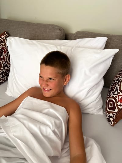 A young male, appearing to be a pre-teen, is depicted in an indoor setting, possibly a residence in Michamvi Kae, Tanzania. He is positioned in a supine or reclined posture, partially covered by white bedding. His head rests against white pillows, which are backed by a textured grey sofa or headboard. The subject has short brown hair and is smiling, looking towards the viewer's right with crinkled eyes. His bare shoulders and upper torso are visible above the white sheet that covers his lower body. A patterned throw pillow with geometric designs in brown, white, and reddish-orange is partially visible on the viewer's left, alongside the white pillow. Another similarly patterned pillow is partially visible on the viewer's right. The background consists of a plain, light-colored wall.