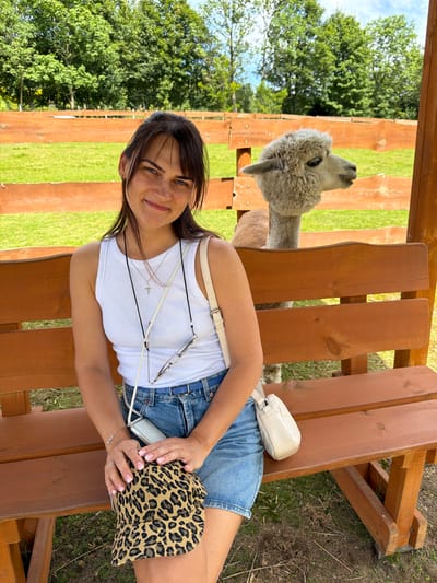 A young woman with dark hair and blue eyes is sitting on a wooden bench outdoors. She is wearing a white tank top, denim shorts, and a leopard-print baseball cap. A small white purse is slung over her shoulder, and she is holding a phone in her hand. Behind her, an alpaca is visible in the background, peeking over a wooden fence. The scene appears to be a farm or an animal sanctuary, possibly in Świeradów-Zdrój, Poland, given the presence of lush greenery and a rustic setting. The woman is smiling and looking towards the camera. The lighting suggests it is daytime, likely a sunny day.